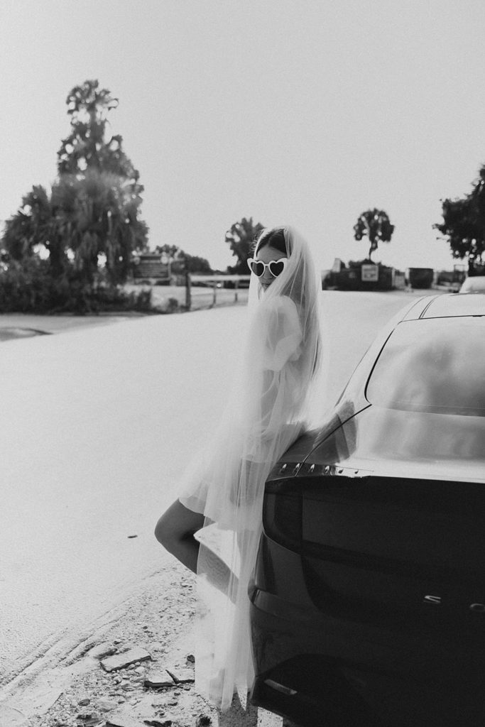 Bride in sunglasses with veil leaning on car
