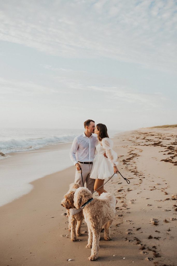 Bride and groom kissing while walking golden doodle dogs on beach