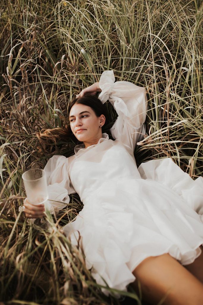 Bride laying in tall beach grass with glass of champagne