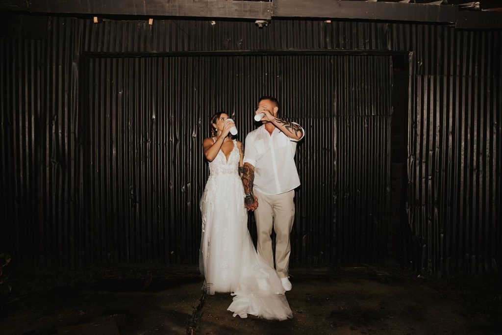 Bride and groom drinking champagne out of cups in front of black wall