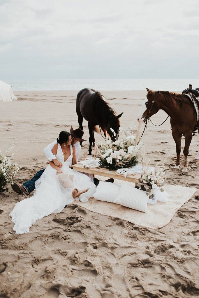 Luxury picnic on Florida beach with pillow seats and floral cake meadow with bride and groom sitting on the ground with their horses