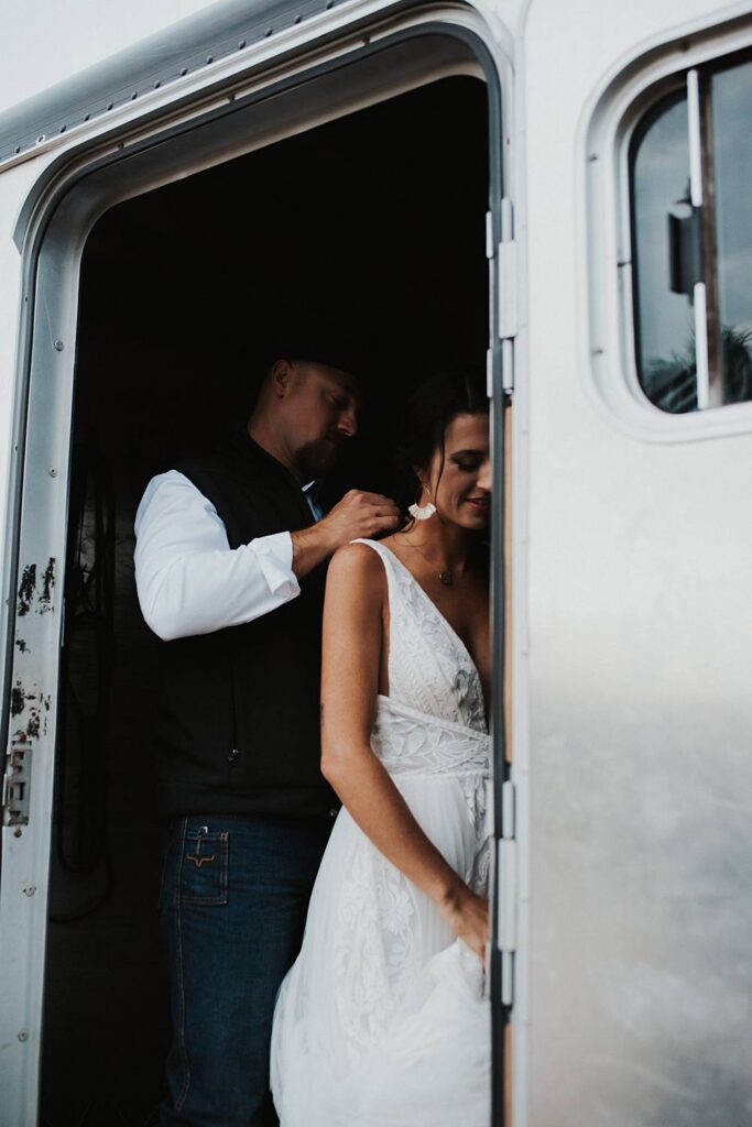 Groom helping bride put on a necklace inside of a horse trailer